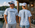 Man and woman wearing Hookset Outdoors Blue Ice camouflage performance fishing shirts on a lakeside dock showing front and back logo designs.