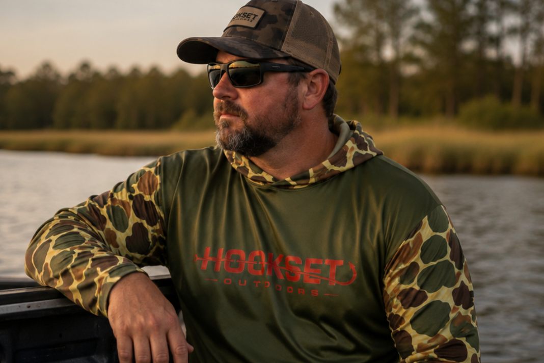 Man wearing Hookset Outdoors Mallard camo UPF 50 fishing hoodie and sunglasses sitting on a boat at sunset