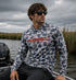 Man wearing Hookset Storm Grey UPF 50 fishing hoodie sitting on a boat in marsh water.