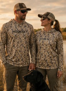 Couple wearing HOOKSET Timber Duck camouflage fishing hoodies with black retriever dog near marsh grass at sunset.